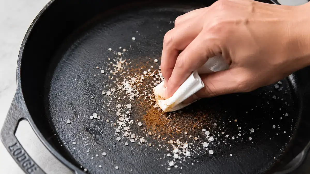 Orange rust spot on the interior of a cast iron skillet next to a scrub brush and coarse salt, before cleaning